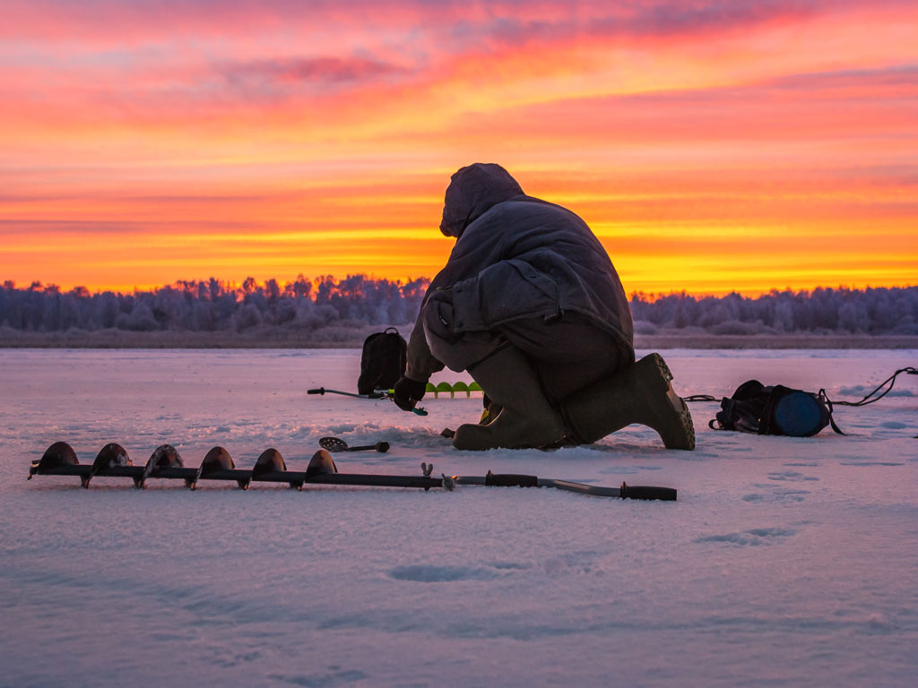 A sunset photo of a man kneeling on a frozen lake, with an auger and other ice fishing equipment spread around him.