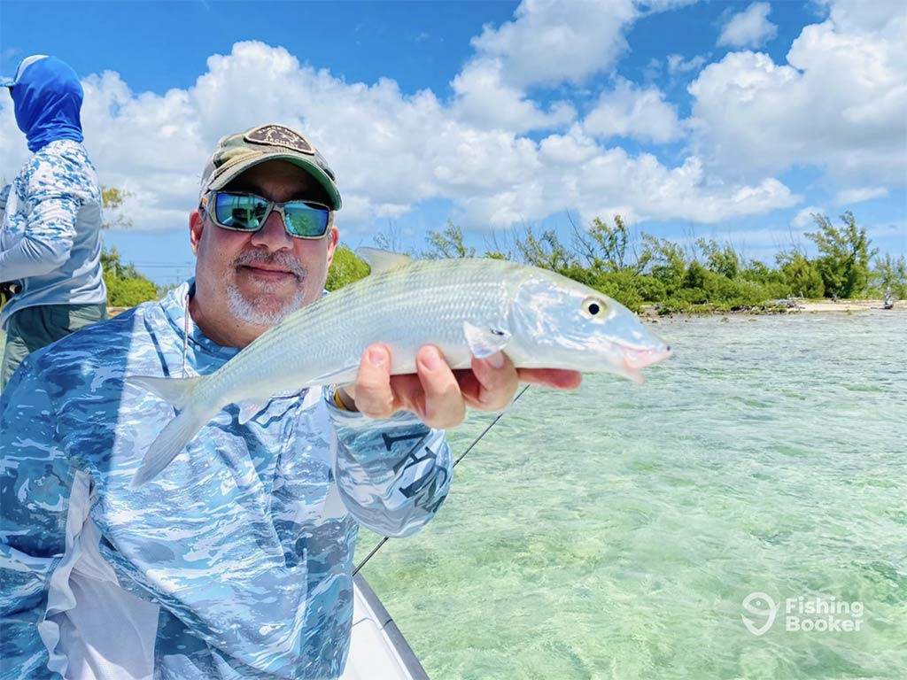 A man in a blue camouflage shirt holding a Bonefish caught in the Bahama aboard a fishing charter, with clear waters around him and some mangrove trees behind him, along with another person in the same outfit standing up on the boat