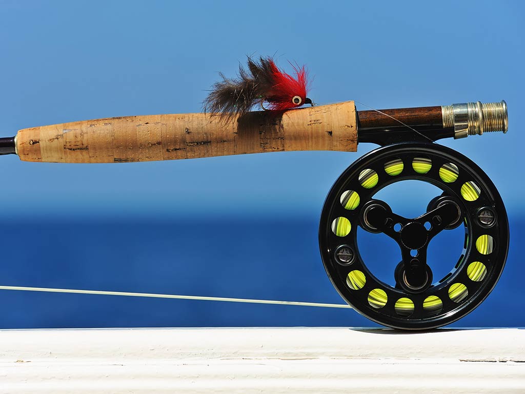 A closeup of a fly fishing reel on a rod, leaning on the side of a saltwater fishing boat on a sunny day with the water behind it