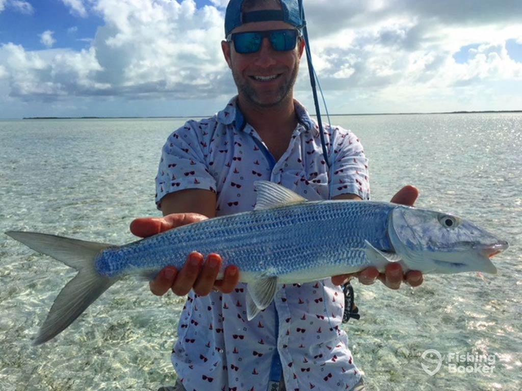 A man in a back-to-front baseball cap and sunglasses holding a large Bonefish to the camera while standing in the shallow waters of the Bahamas on a day with sunny intervals