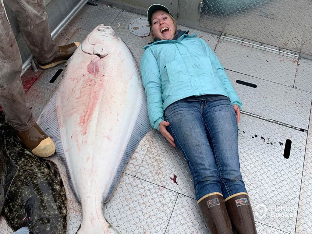 A woman lying down on the floor of a fishing boat in Seward, Alaska, next to a Halibut splattered with blood that's just as tall as her
