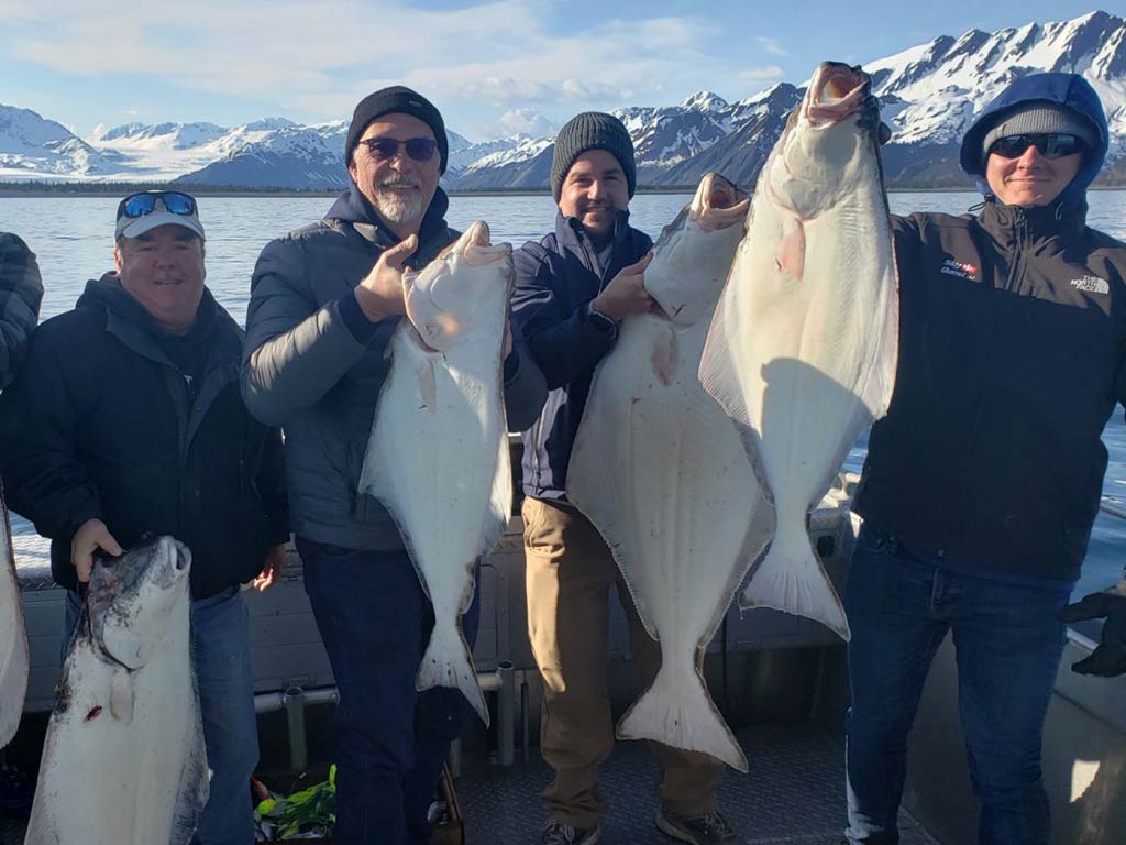 Four men stood on a fishing charter in Seward, Alaska, each holding a Halibut, with snow-capped mountains visible across the water in the distance