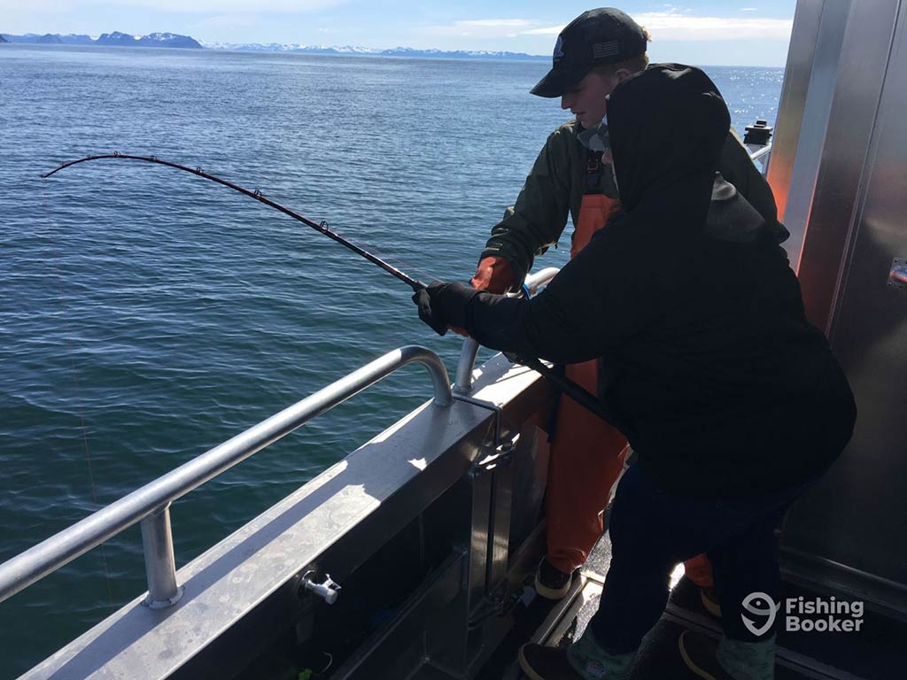 One angler helping another with a heavy-action fishing rod aboard a charter boat in Seward, Alaska, on a clear day, as they try fishing the deep waters