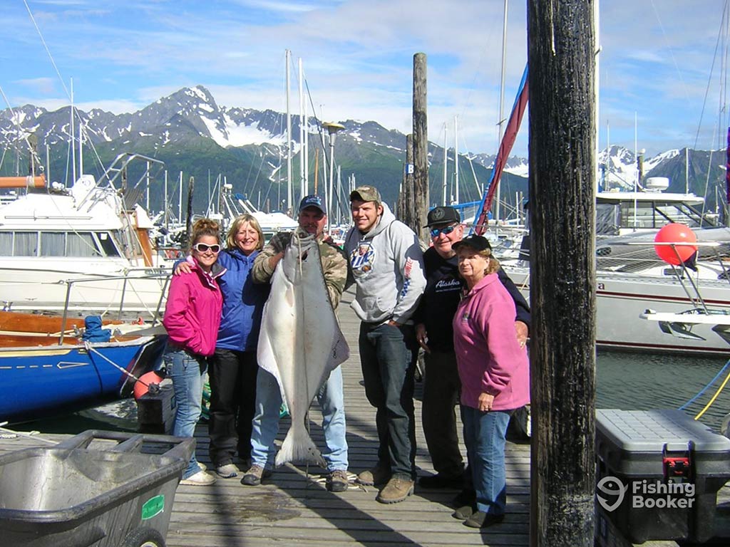 A group of three men and three women on a boat dock in Seward, Alaska, posing with one large Halibut while surrounded by boats and with snow-capped mountains visible behind them on a clear day