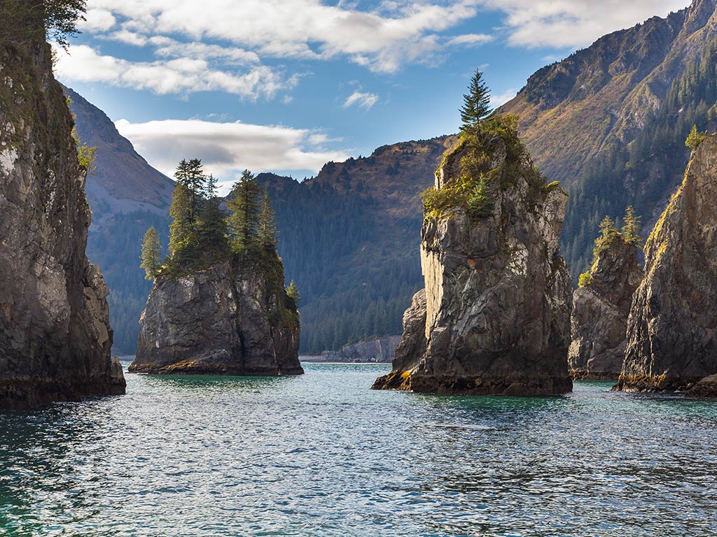 A view across the water towards some impressive rock formations sticking out of the water in Resurrection Bay near Seward, Alaska, on a day with sunny intervals in summer