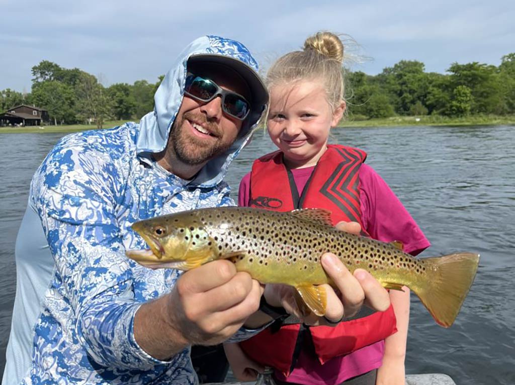 A man in a hood and sunglasses kneeling next to a little girl while holding a sizeable Brown Trout with open waters visible behind them stretching to a tree-lined shore in the distance