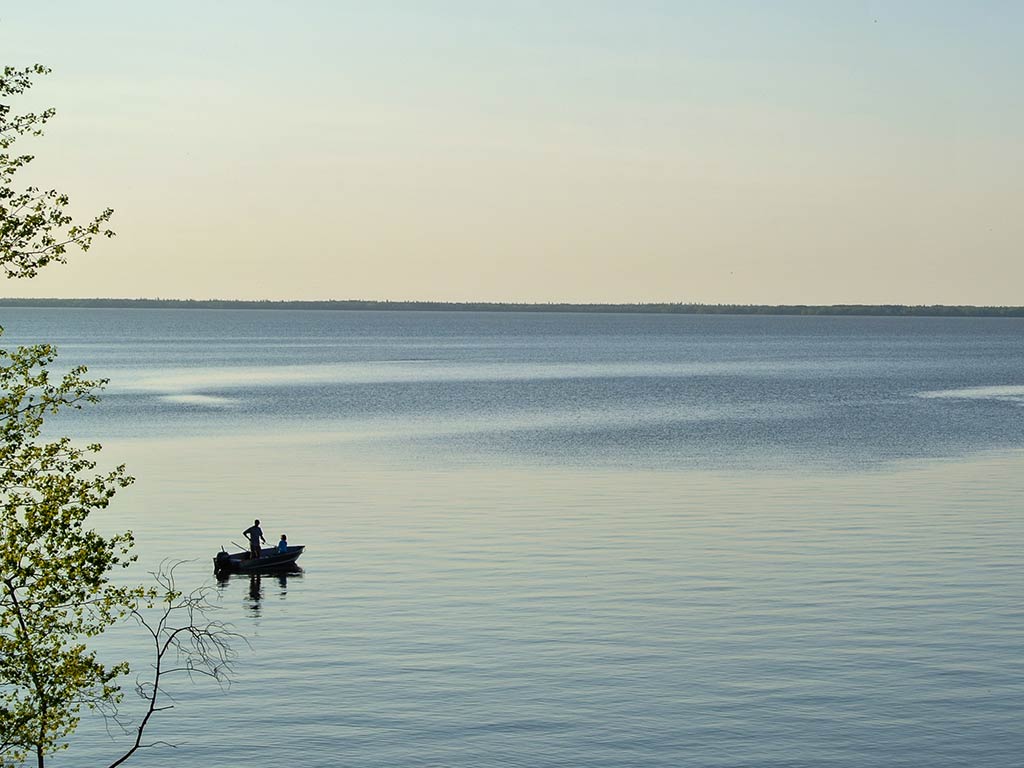 A view from a hill across some trees on the left of the image towards a lone boat featuring an angler on the vast waters of Lake Winnipeg at sunset on a clear day