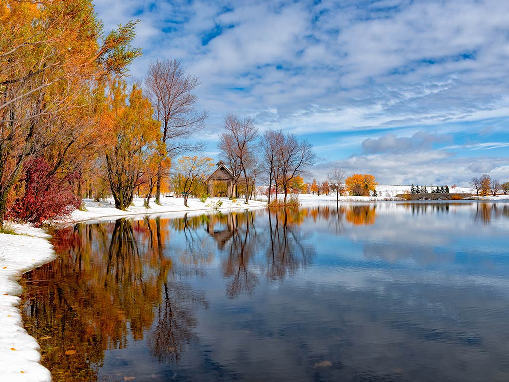 A view across the water towards a snow-covered shoreline of Lake Winnipeg on a clear day, with red-leaved trees visible on the shore