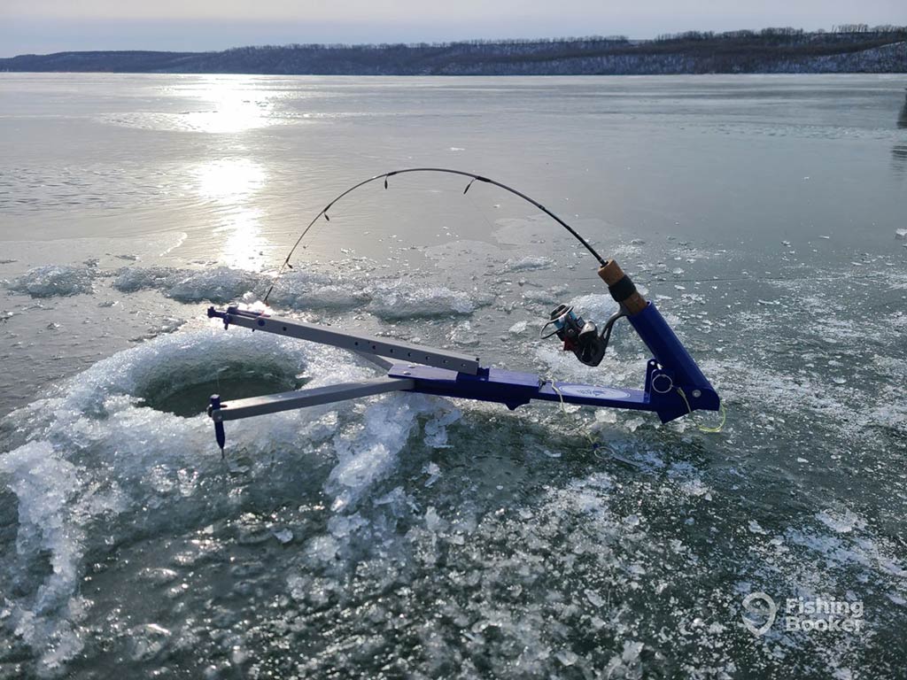 A closeup of a hole in the blue ice of a frozen lake with a small ice fishing rod set up for fishing near sunset on a clear winter's day