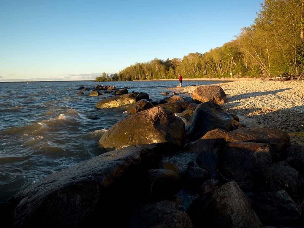 A view along a rocky shoreline of Lake Winnipeg near sunset on a clear day, with a lone angler visible on a beach in the distance in front of some alpine trees