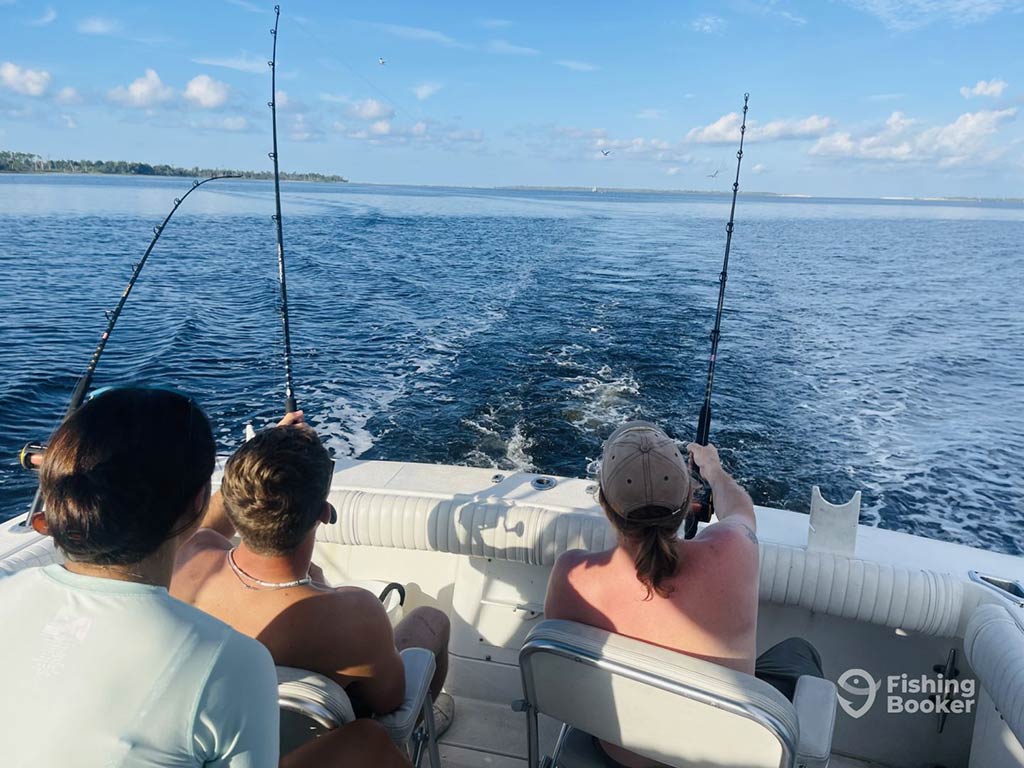 A view across the deck of a fishing charter in Panama City, Fl, with three men sitting and enjoying the ride while three trolling rods trail lines in the boat's wake on a clear day