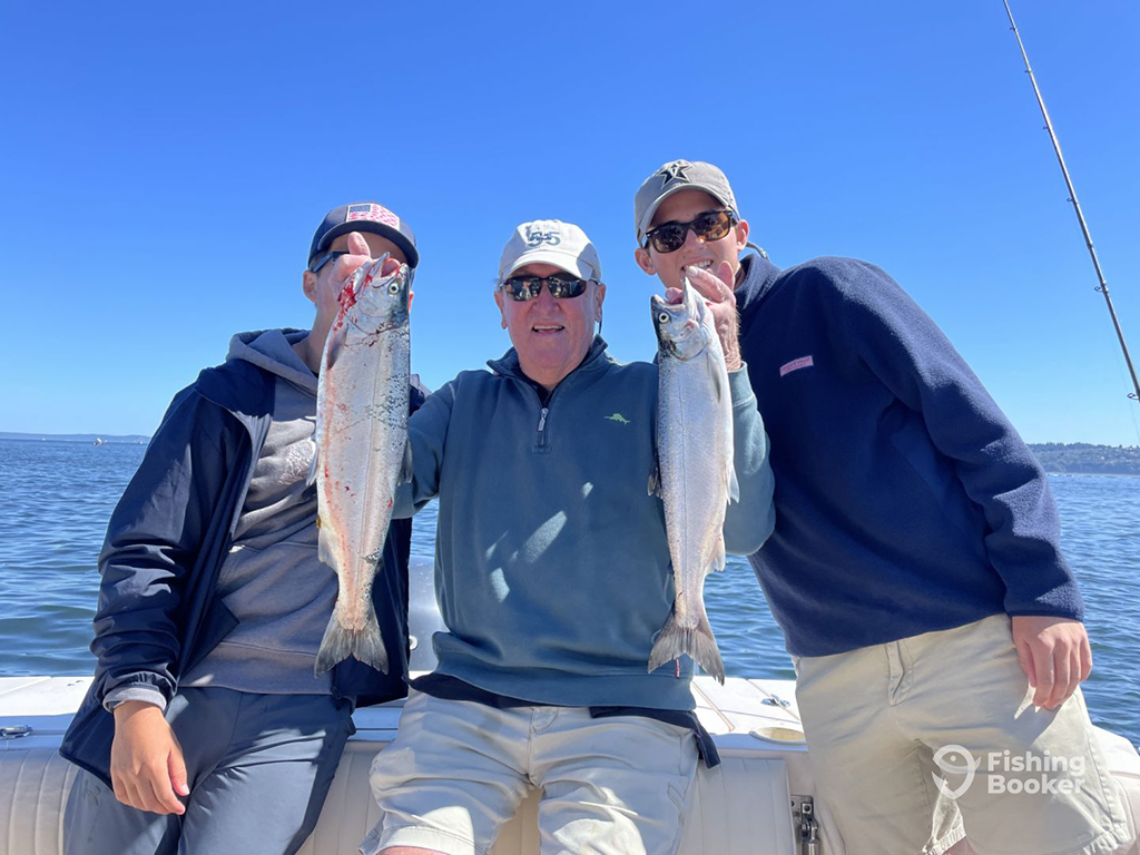 Three anglers aboard a boat on a lake on a sunny day, with the angler in the center of the image holding two Coho Salmon