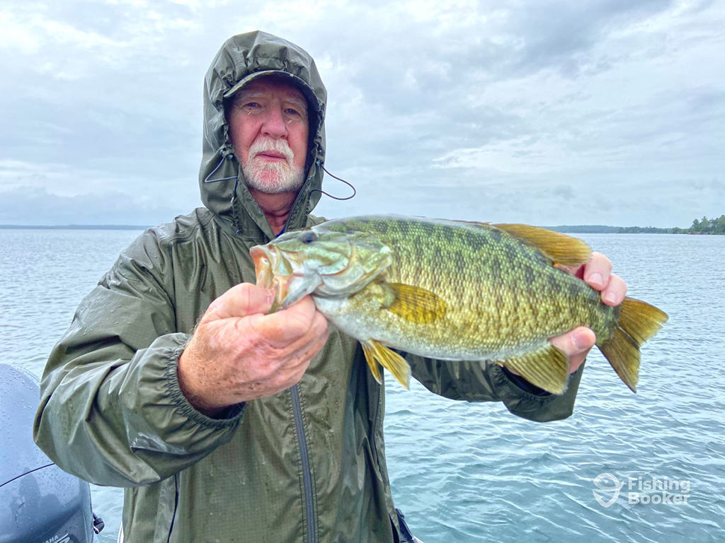 An angler in a raincoat on a lake on a cloudy day holding a large Smallmouth Bass with the boat's motor and water in the background