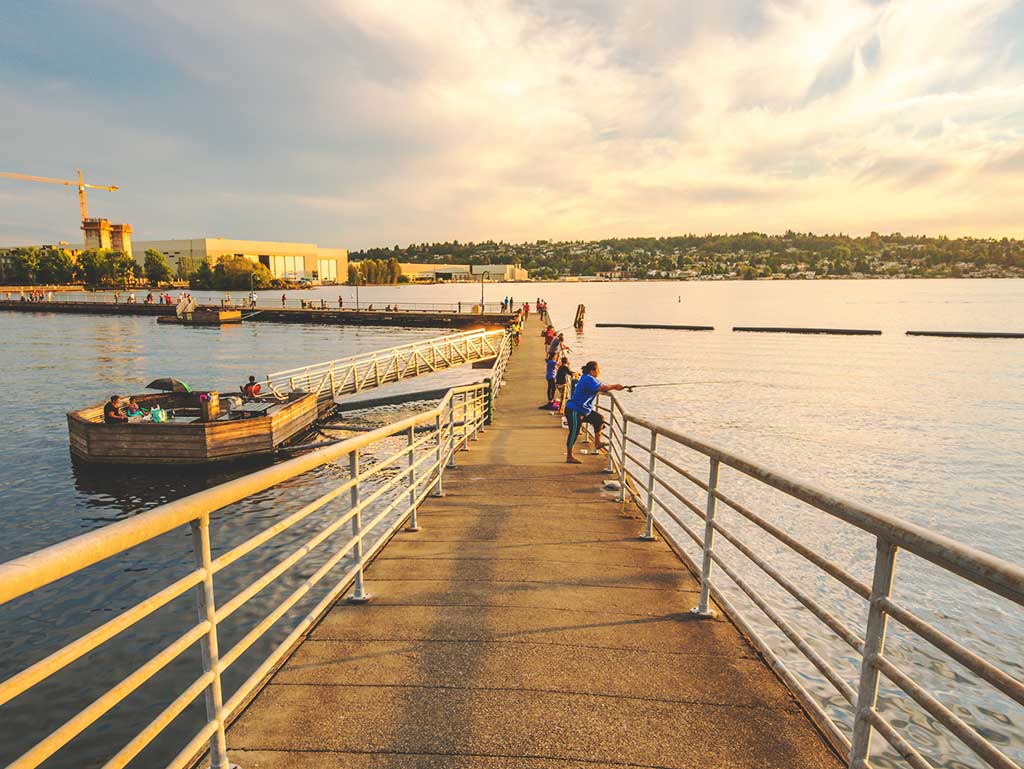 A view along a fishing pier and dock towards a woman casting a line on Lake Washington as the sun sets against an industrial background