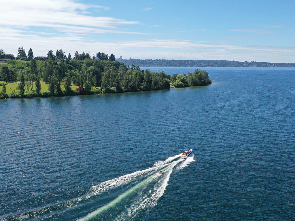 An aerial view of a single motor boat crossing Lake Washington on a sunny day next to a grassy shoreline