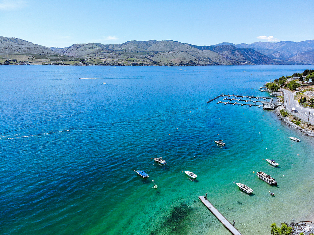 An aerial view of clear blue green waters of Lake Chelan on a sunny day with small boats moored in the foreground and small mountains in the background on the other side of the lake