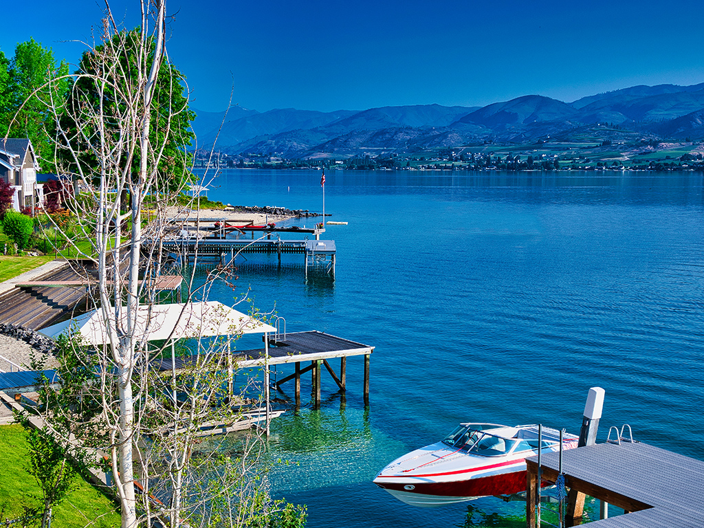 A view from a residential shoreline of Lake Chelan with docks stretching into the lake and a single boat moored at the front of the photo on clear blue water on a sunny day with mountains in the backdrop