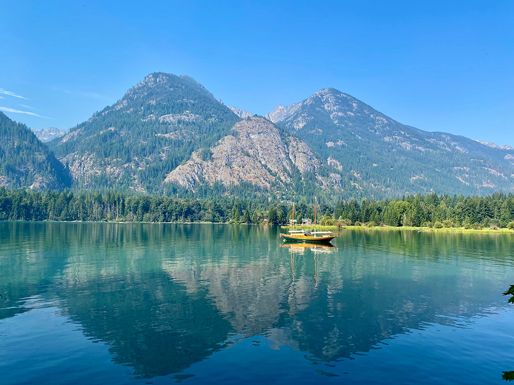 A single yellow boat floating on the lake in the middle of the photo, against a forested mountainous backdrop, with the clear water reflecting the boat and pointy mountains