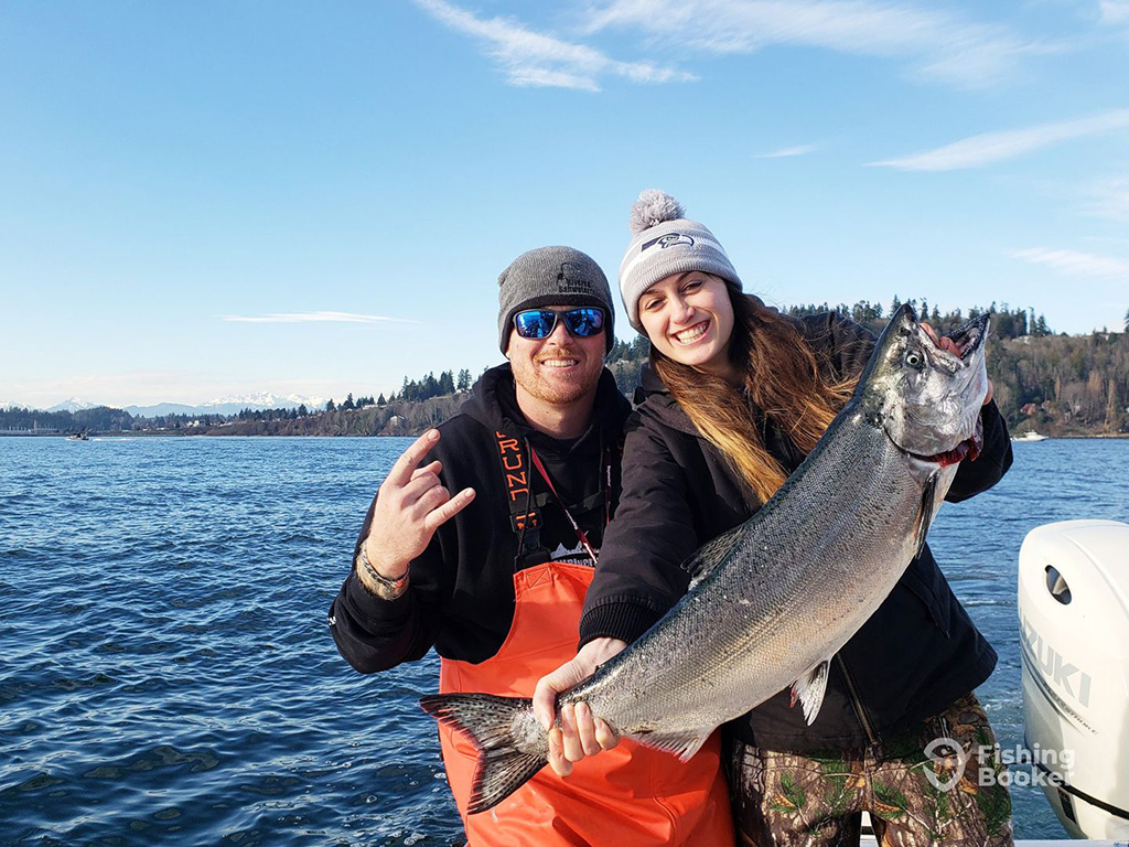 A fishing guide in orange overalls standing jubilantly next to a happy woman holding a Salmon on a boat on a sunny day 