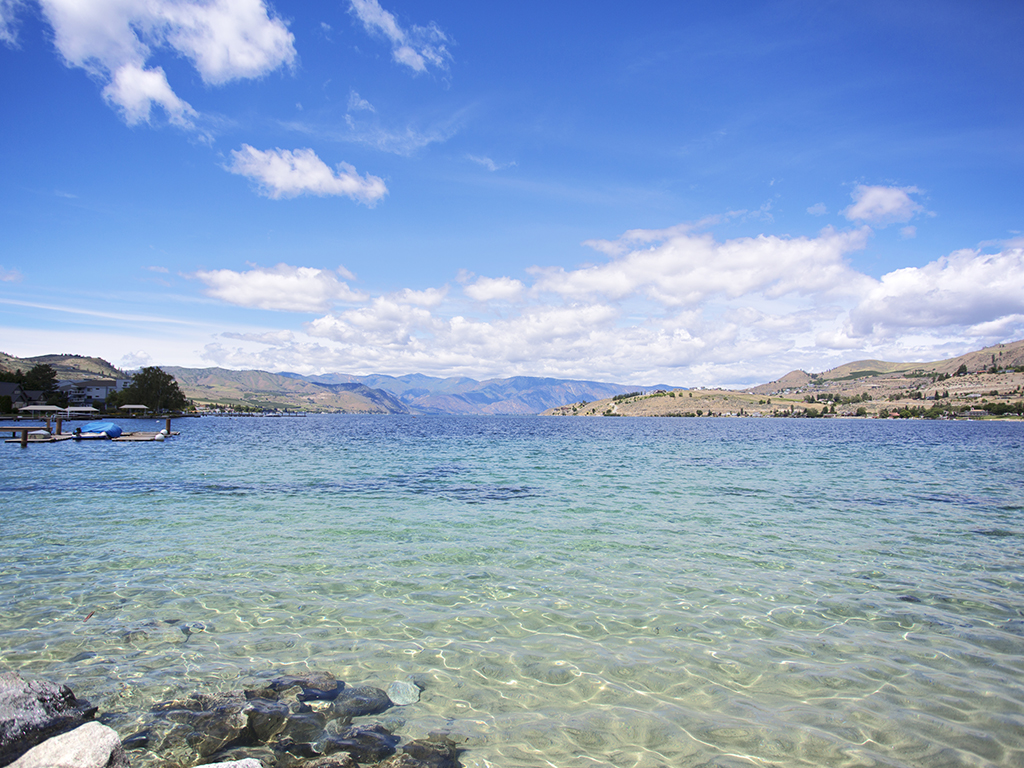 A view of the very clear, shallow waters of Lake Chelan on a sunny day with rocky shoreline visible to the left of the photo, a dock further away, and mountains in the distance