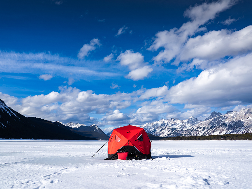 A solitary ice fishing tent pitched on a frozen lake in Canada amongst some snowy mountains on a sunny day