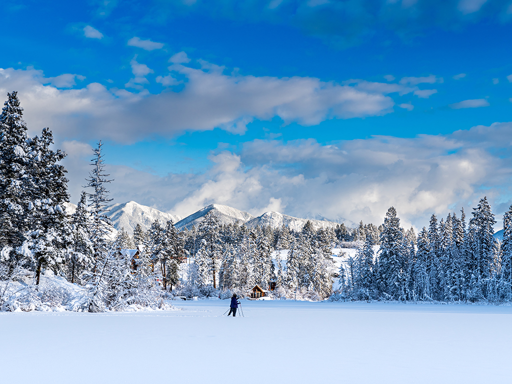 Two people cross a frozen lake in the Kootenay Rockies near Golden, BC, with snowy pine trees and white mountains in the background