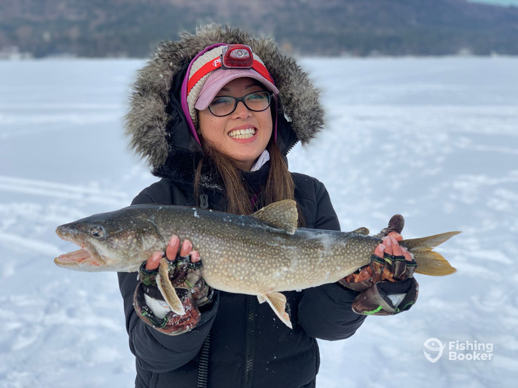 A female angler in full winter gear standing on a frozen lake holding a Lake Trout that she caught while ice fishing in Canada