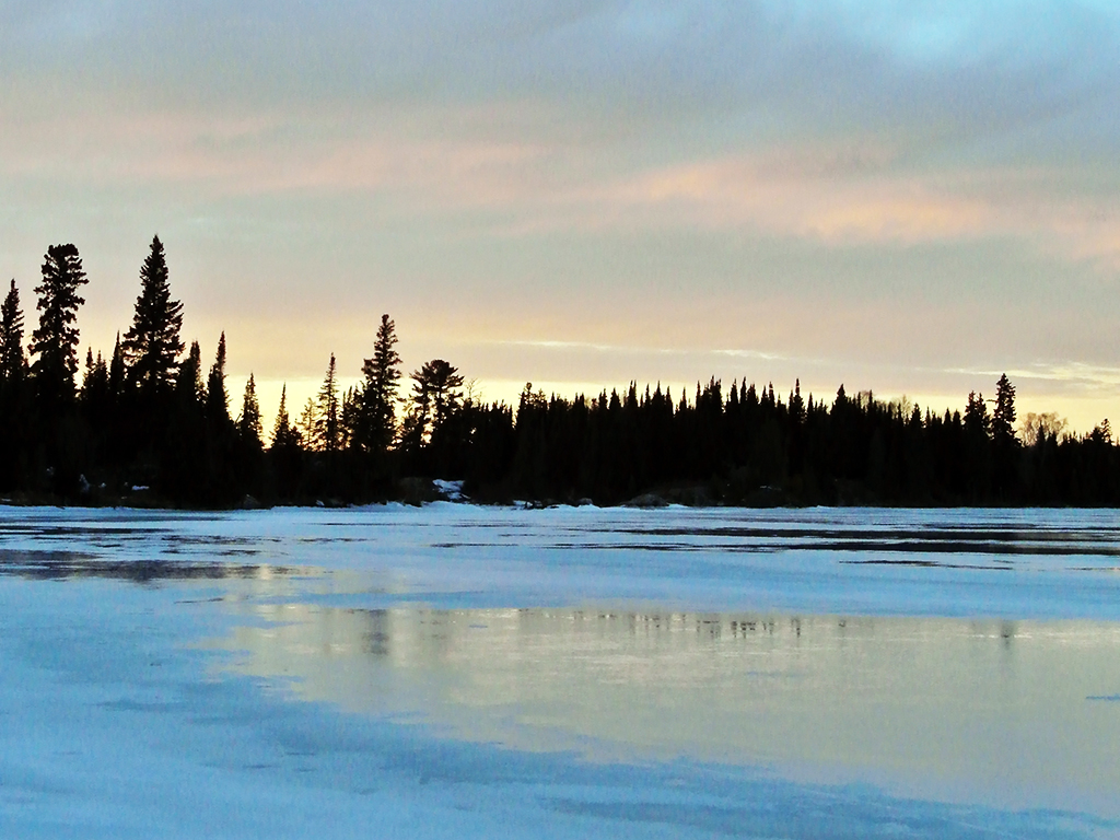 A view across the frozen Falcon Lake at dawn, with a line of trees reflecting in parts of melting ice