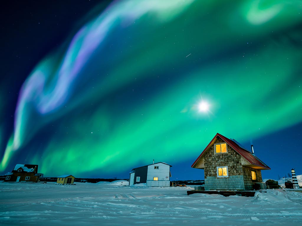 A view across snow-covered terrain twards loosely gathered houses near the Great Slave Lake, with the Northern Lights in full view above.