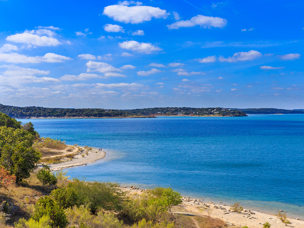 A view of the bright blue waters of Canyon Lake on a sunny day with a tree-lined sandy shoreline to the left of the photo