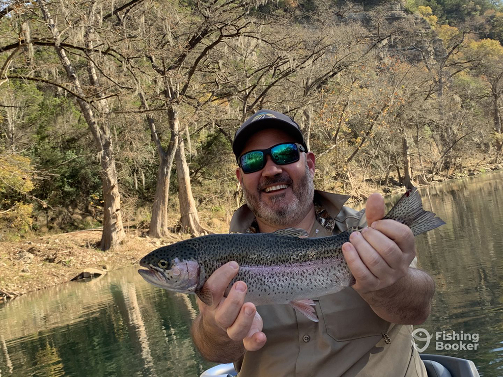 A smiling man in sunglasses holds a Rainbow Trout on the lower Guadalupe River by a tree-lined bank on a sunny day in the winter