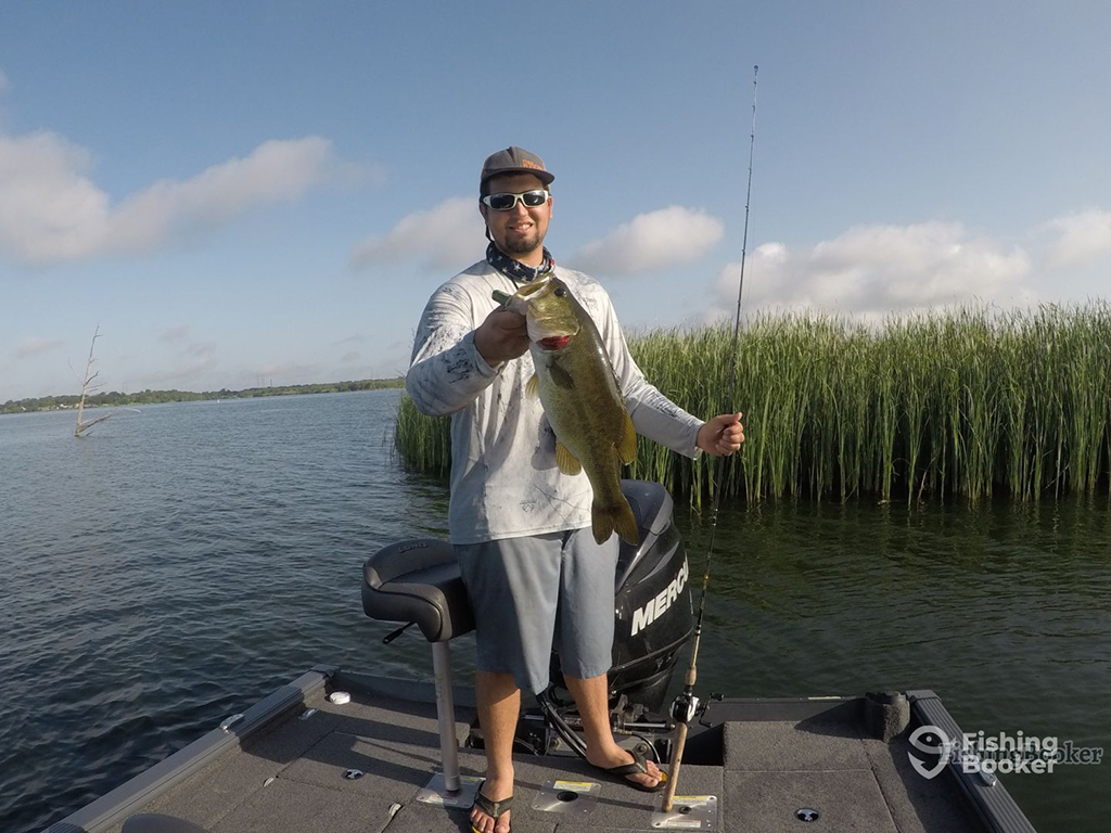 A smiling man stands on a Bass boat holding a large Smallmouth Bass in one hand and a fishing rod in the other next to a clump of reeds