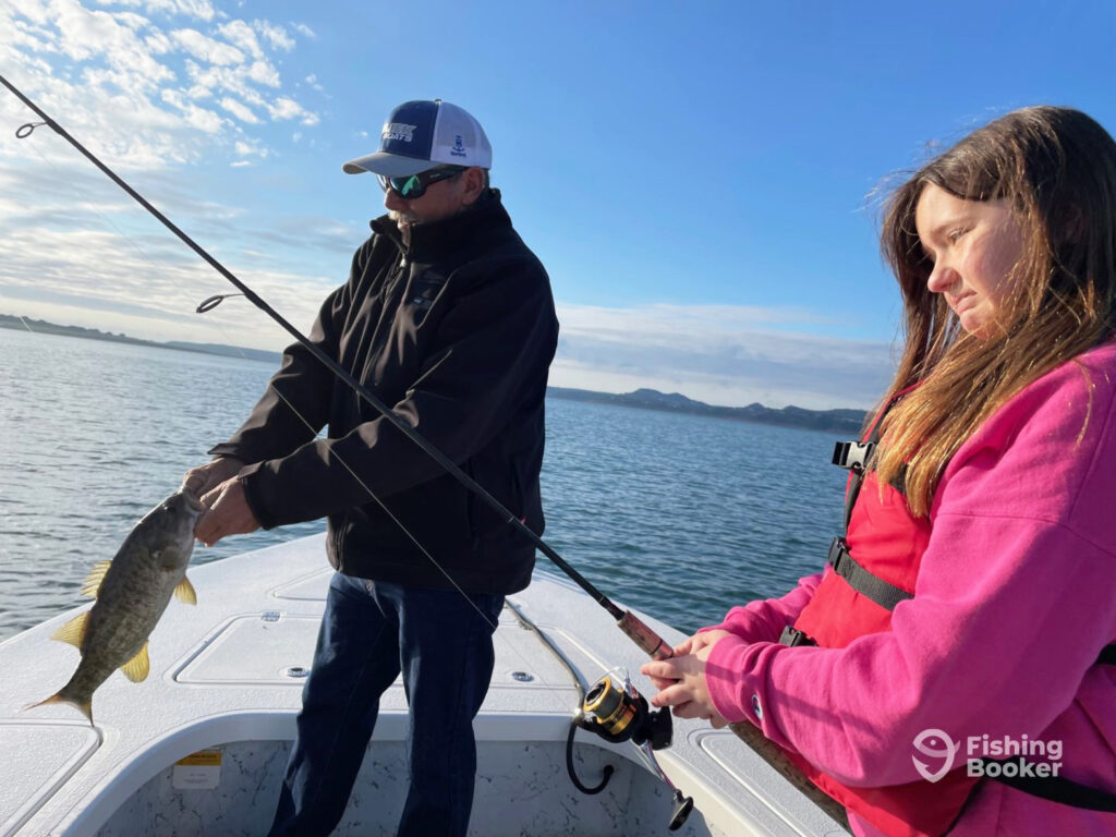 A young girl stands on a boat holding a fishing rod while a Canyon Lake fishing guide removes a Smallmouth Bass from a hook at the end of her line on a sunny day in winter