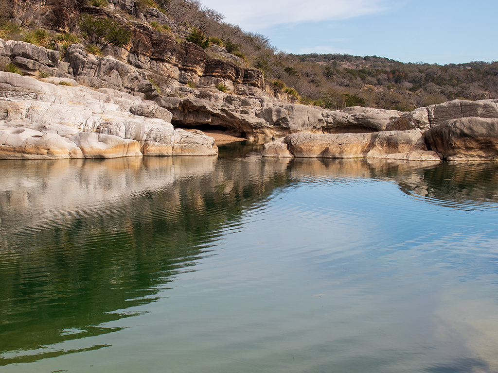 A view across the calm waters of Canyon Lake towards limestone cliffs in Texas Hill Country, mirrored in the clear waters of 