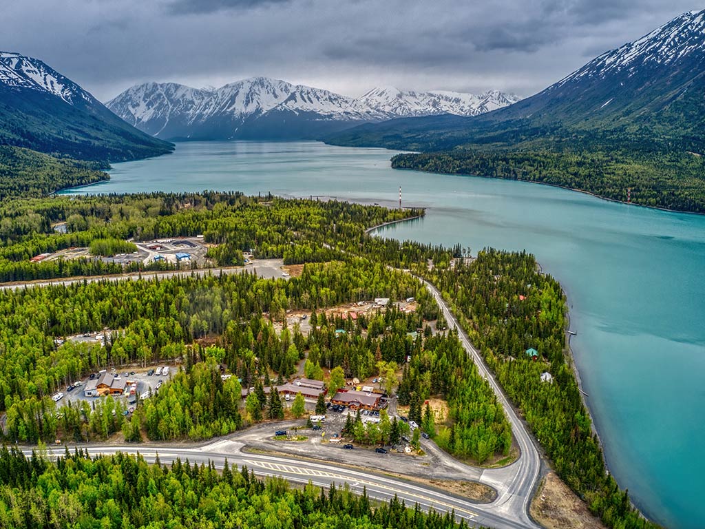 An aerial view of Cooper Landing, Alaska, on a cloudy day with crystal clear waters on the right of the image, the town on the left and snow-capped mountains in the distance