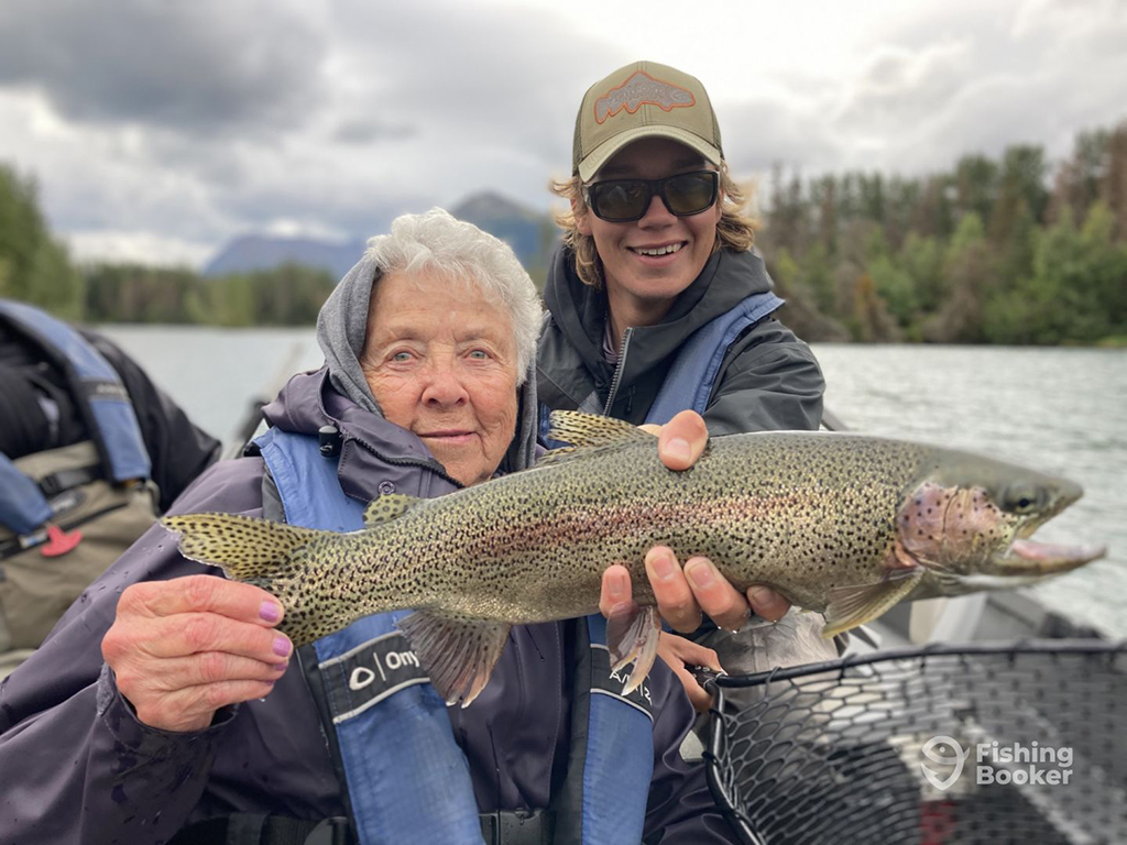 A fishing guide and an older female angler hold a Rainbow Trout aboard a drift boat on a river with a release net visible and pine trees and mountains in the distance