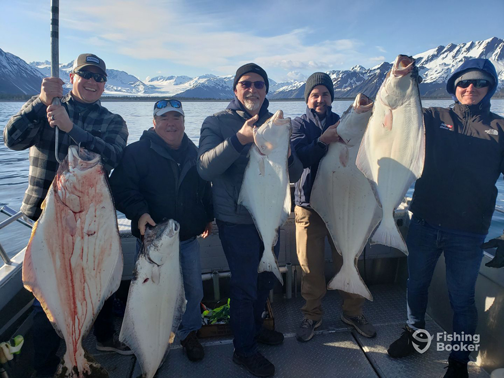 Five anglers on a charter boat hold a Halibut each against a backdrop of snow-covered mountains on a clear day