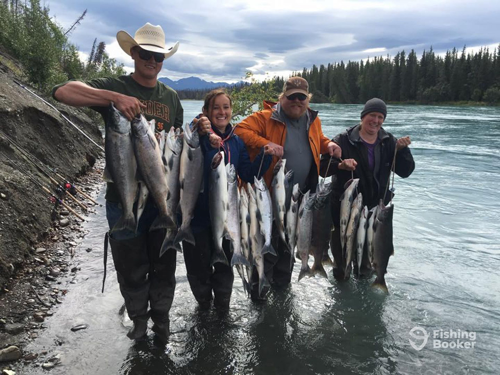 Four anglers in waders hold a string of Salmon with their fishing rods visible leaning against the river bank on a cloudy day in Alaska