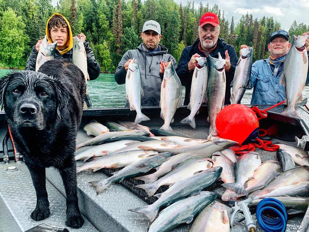 A group of four anglers holds a large string of Salmon behind a boat that is filled with more Salmon in Cooper Landing with a wet dog standing to the left of the group