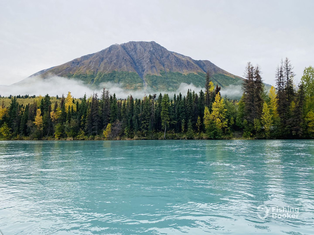 A rocky mountain looms above the cloud across the bank from the blue green river in the foreground in Cooper Landing