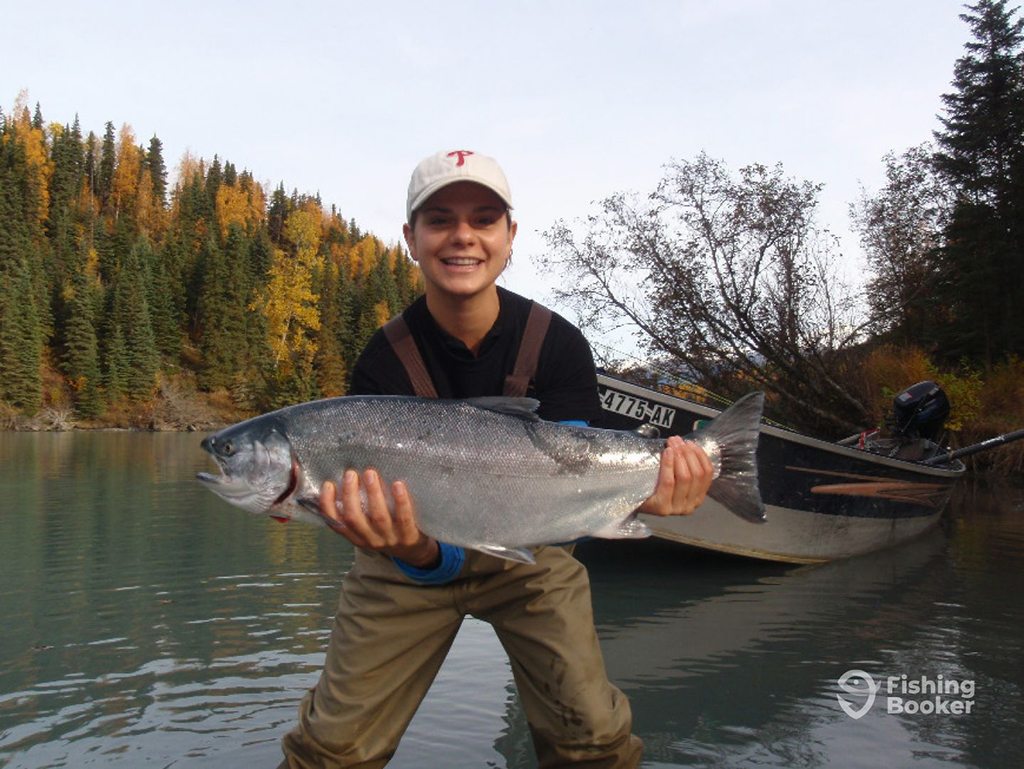 A female angler in waders stands in a river in Cooper Landing next to a drift boat holding a large Salmon against fall colors in the background