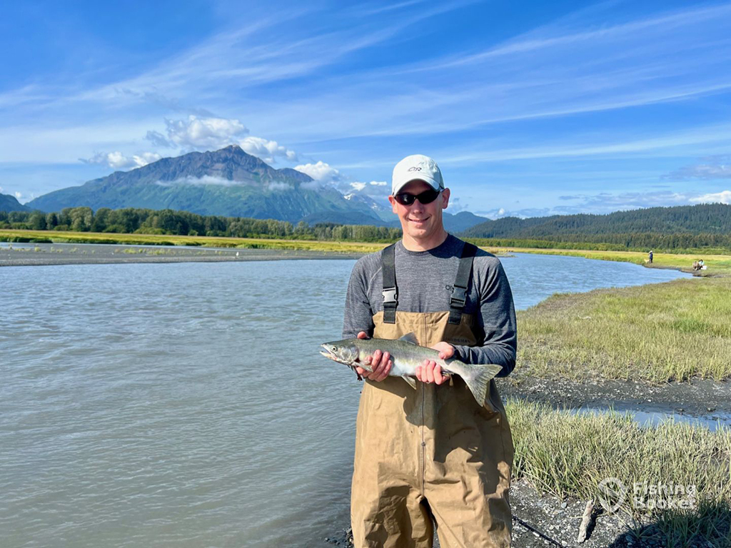 A male angler in waders holds a small Trout next to a river in Cooper Landing, AK, on a sunny day with grassy river banks and a mountain in the background