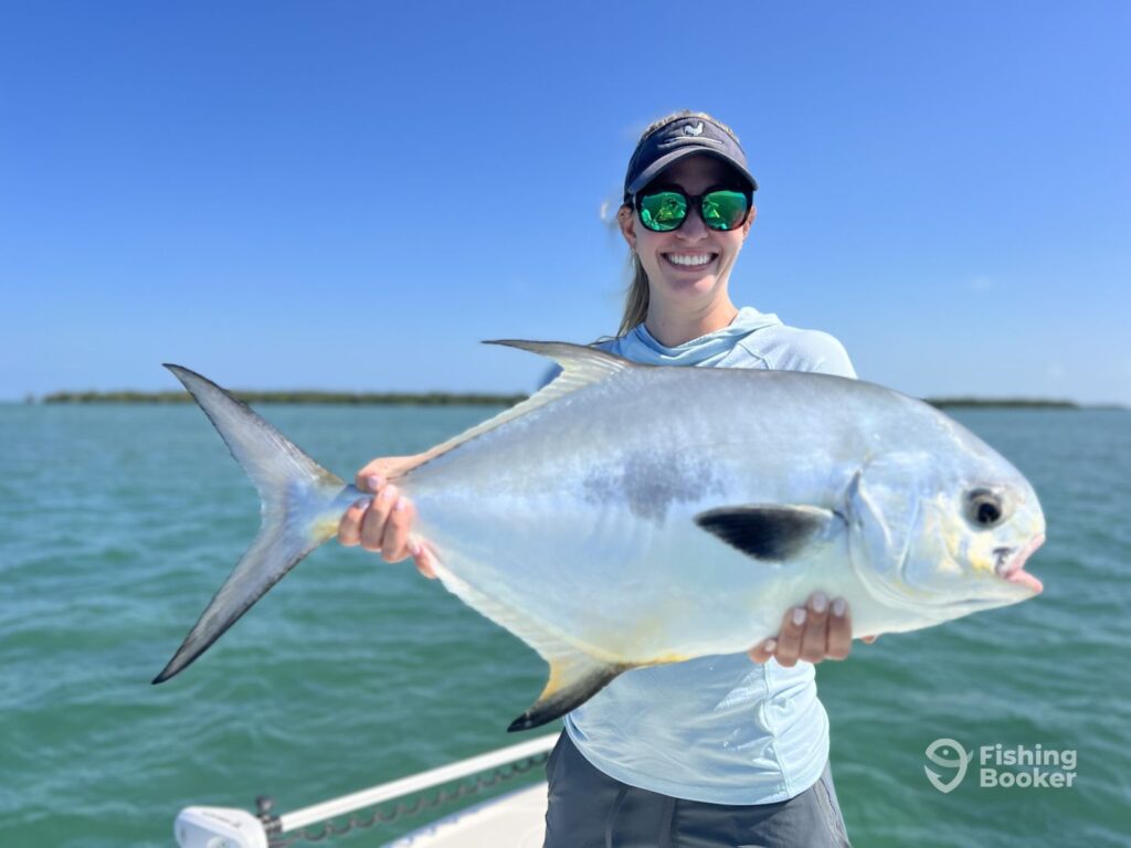 A female angler holds a large Permit on a boat on the flats in Key West with a trolling motor visible behind her on a sunny day