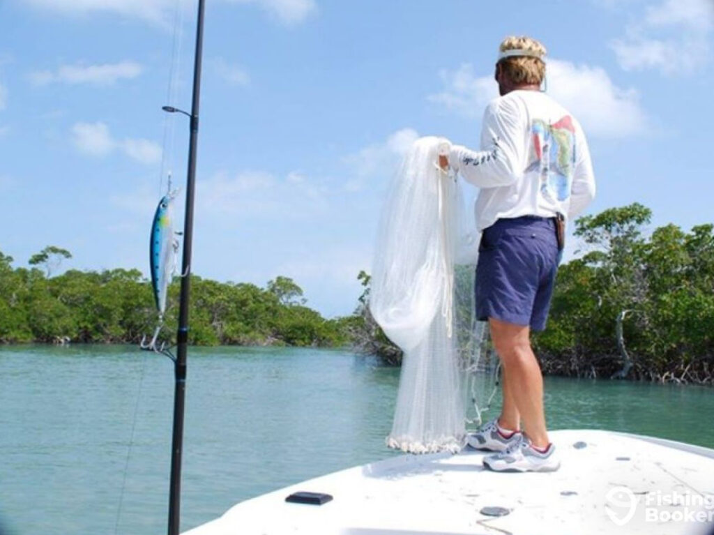 A charter captain in Key West prepares to throw a cast net to catch live bait near some mangroves. An artificial lure is visible attached to a fishing rod to the left of the photo