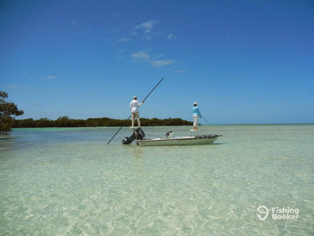 A charter captain stands atop a poling platform as he directs his skiff across the flats of Key West with a female angler standing at the front of the boat and fishing from a casting platform on a clear day