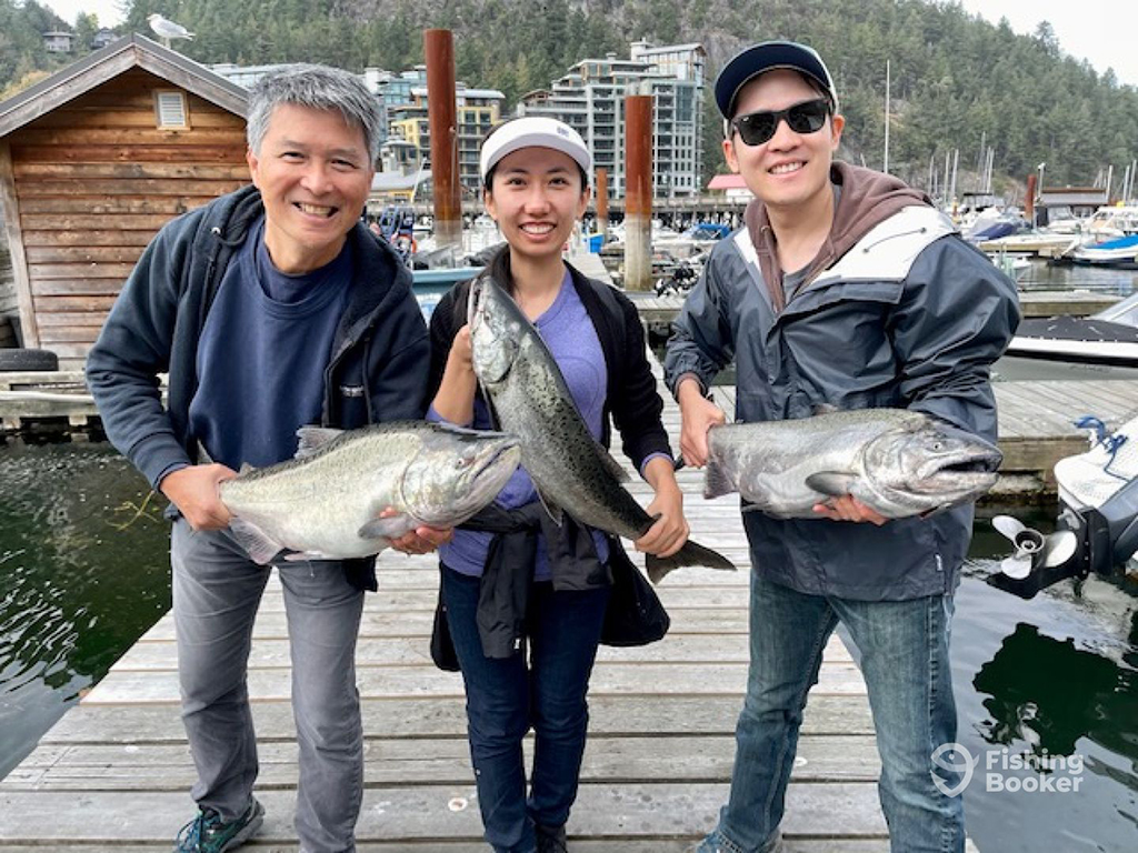 A group of three anglers holding a large Salmon each on an urban wooden dock at a marina in Vancouver after a fishing trip on a cloudy day