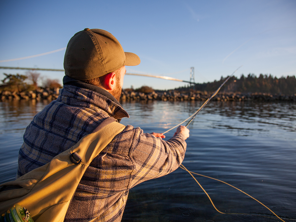 A male angler holding a fly fishing rod out into some water as the sun is setting near a bridge in West Vancouver