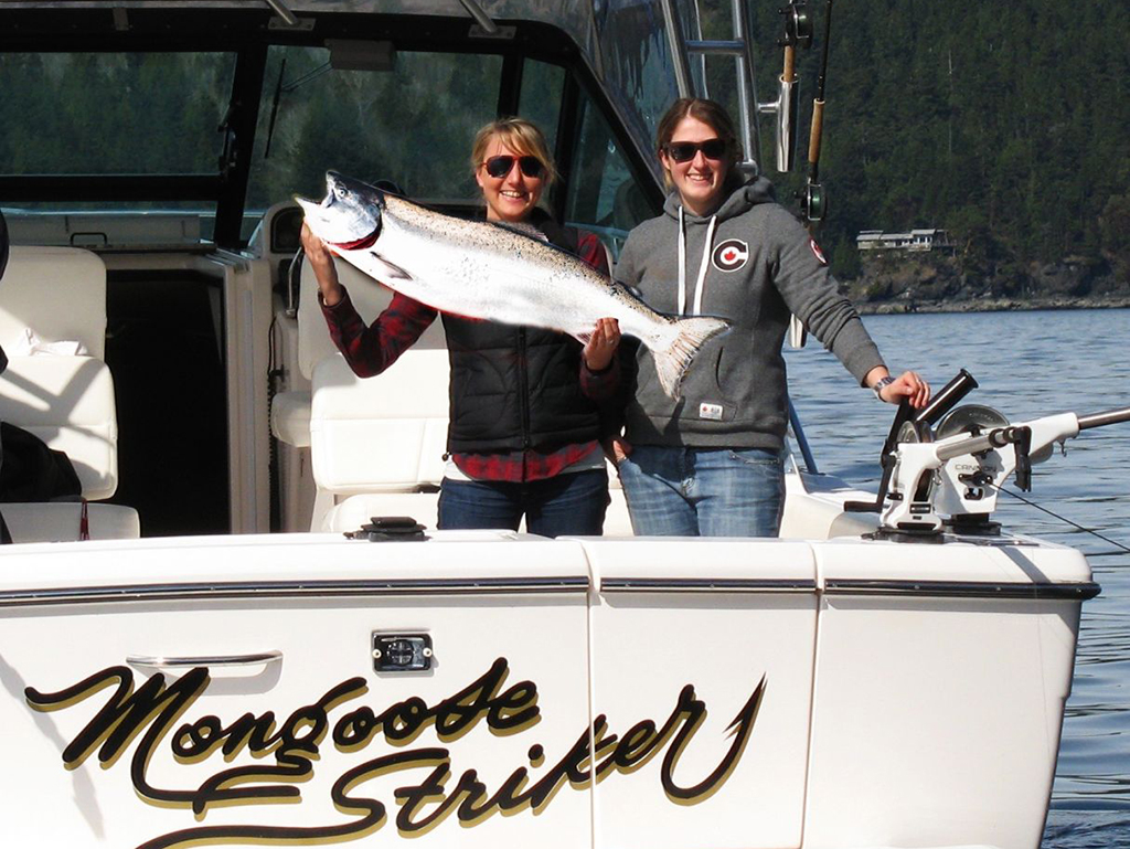 Two female anglers holding a Salmon aboard a fishing charter boat named Mongoose Striker with fishing gear visible on the boat