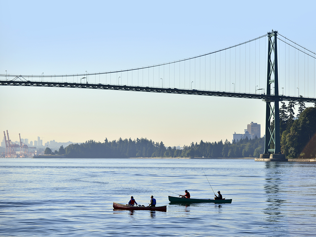 Four anglers fly fishing from two kayaks underneath a Vancouver bridge with the city in the background on a bright, hazy day
