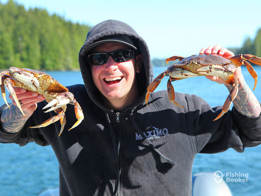 A man smiling as he holds two large Crabs on a cold day in Washington state with a backdrop of blue water and green evergreen trees 